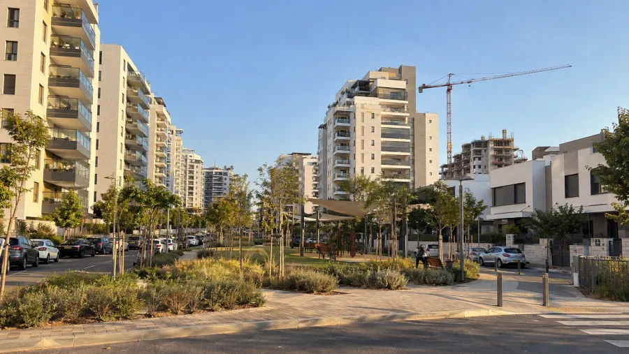 שכונת מגורים חדשה: בנייני מגורים גבוהים עם חיפוי אבן, גינה ציבורית עם עצים צעירים ומנוף בנייה ברקע