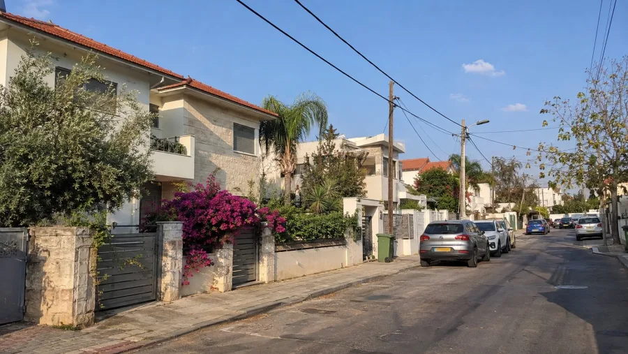 רחוב בשכונת בתים פרטיים ביום שמשי: בתים בני שתי קומות עם חיפוי אבן וגג רעפים אדום, דקל, שיחי בוגנוויליה, עמודי חשמל ורכבים חונים
